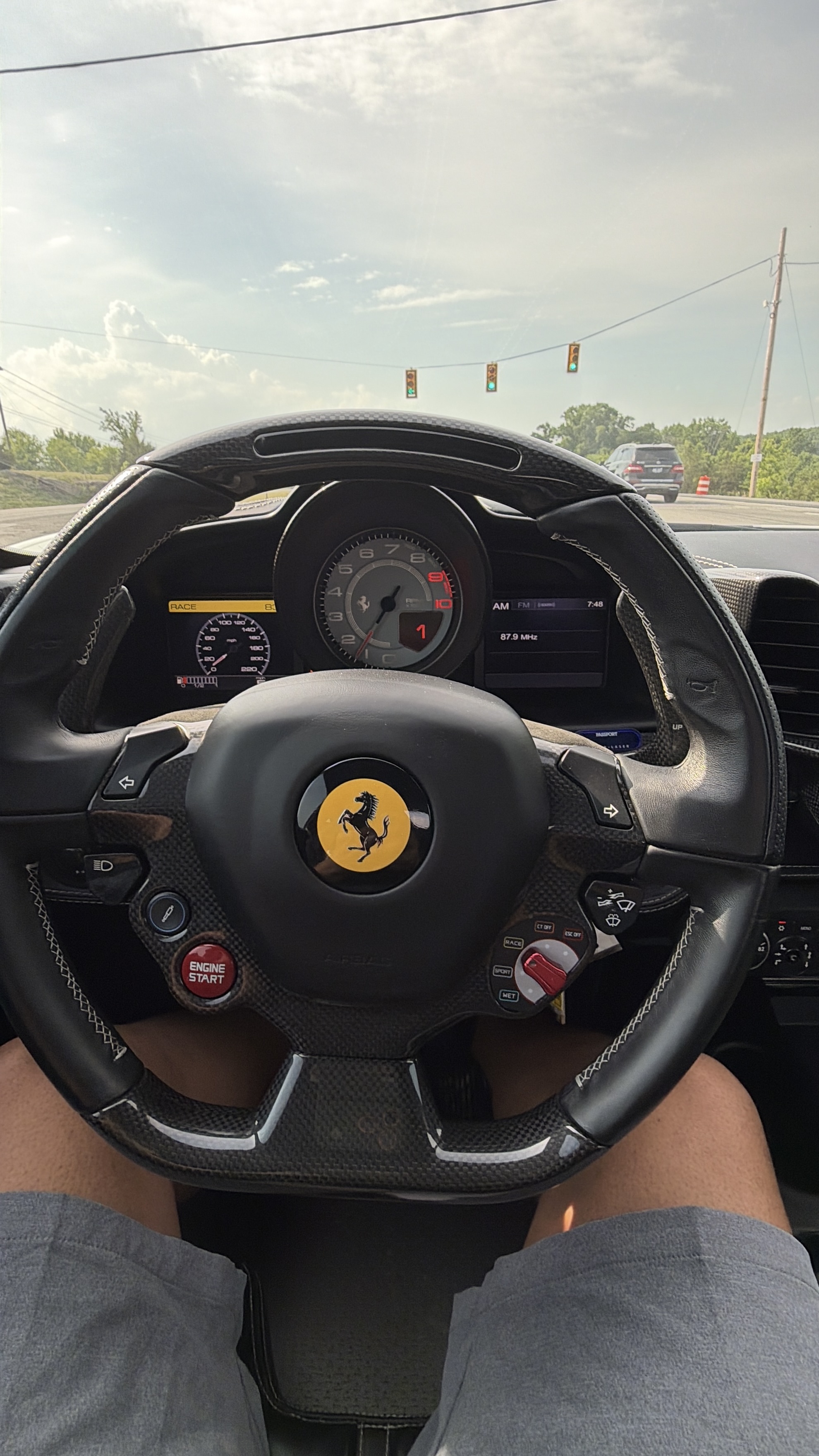 Ferrari cockpit POV — Prancing Horse steering wheel, carbon fiber, ENGINE START button, green lights at intersection