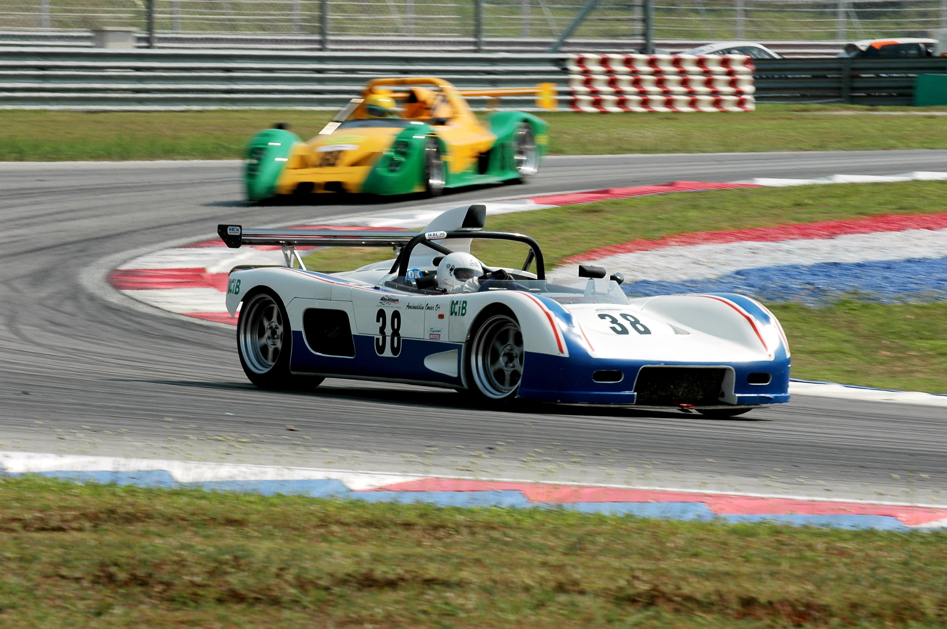 Open prototype race cars cornering on circuit, white blue #38 leads green yellow car